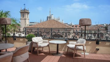 A terrace with wicker tables and chairs, a view of a historic building with a tower and spires, and a wrought iron railing.