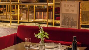 A dining area with a red upholstered booth, a table set with plates, glasses, and a vase with a plant. There is a book titled "IL DUOMO DI MILANO" on the wall behind the booth.