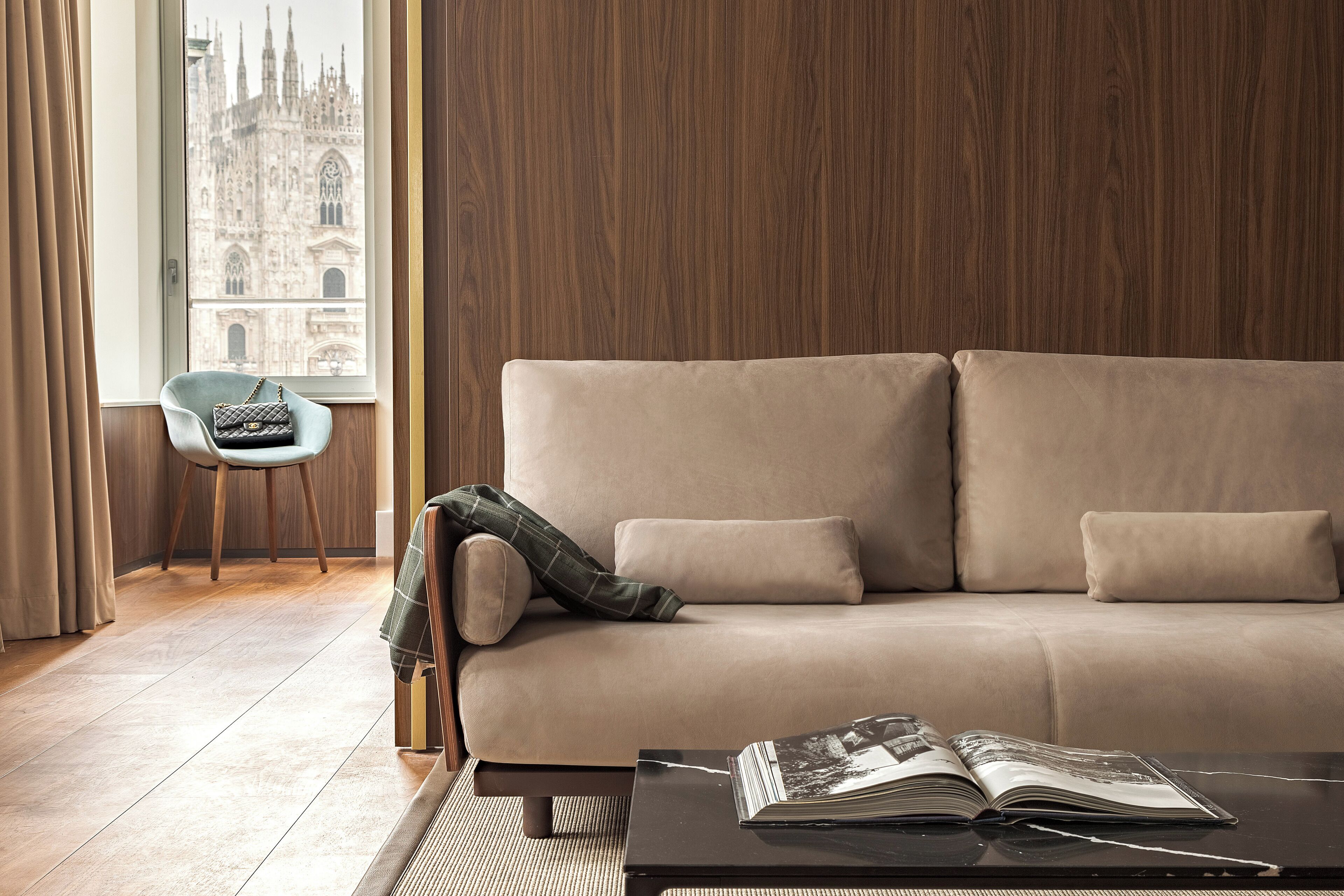 A modern living room with a beige sofa, a small table with a magazine, and a view of a historic building through a window.