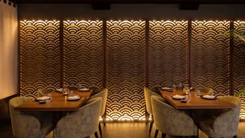 Un coin repas avec une table en bois dressée pour quatre personnes, entourée de chaises rembourrées. Un mur décoratif orné d’un motif ondulé et un éclairage chaleureux se trouvent derrière la table.