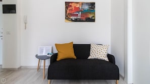 Un salon moderne avec un canapé noir, des coussins jaune moutarde et à pois, une petite table d’appoint sur laquelle est posée une affiche encadrée, et un tableau abstrait coloré accroché au mur.