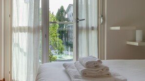 Quarto de hotel com cama, janela com cortinas leves, vista para edifícios e árvores, e uma toalha branca sobre a cama.