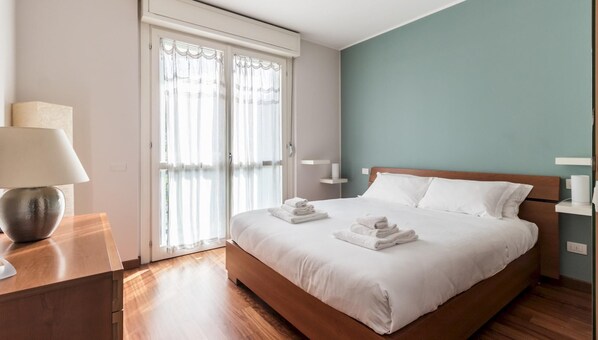 A neatly made bed with white linens and towels, a bedside table with a lamp, and a window with sheer curtains. - Cavour 75 (Cernusco sul Naviglio)
