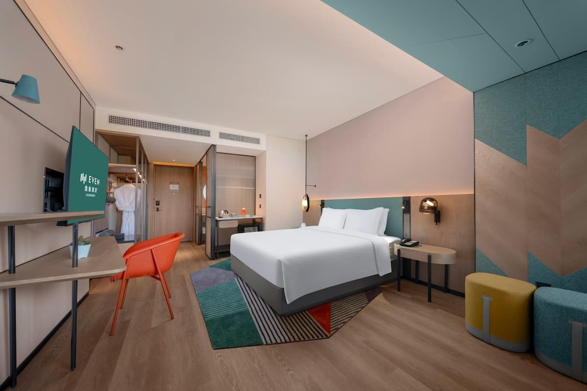 A hotel room with a bed, desk, chair, and a small table with a lamp.