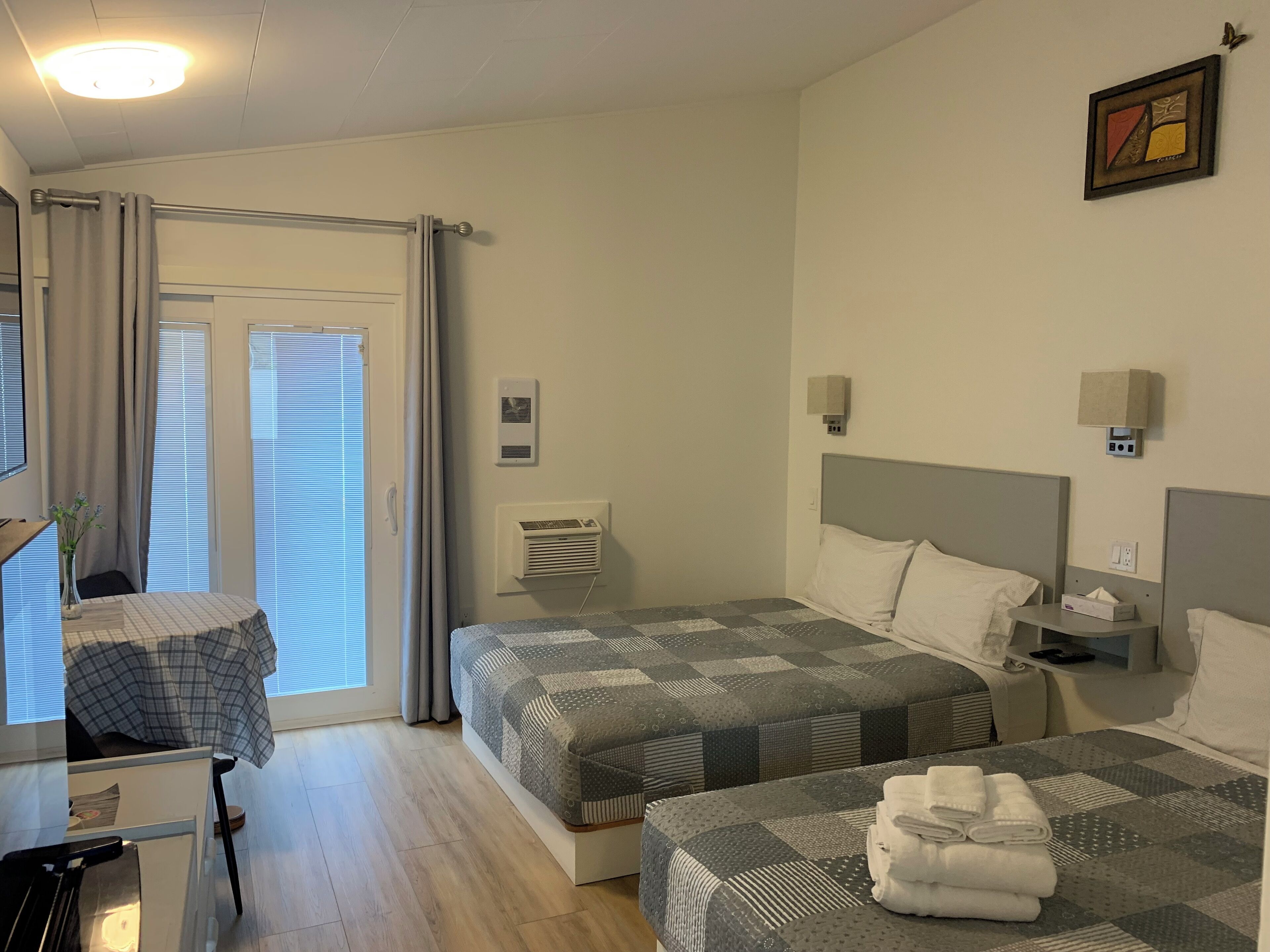 A hotel room with two beds, a desk, a chair, a window with curtains, an air conditioning unit, and a bedside table with a lamp.