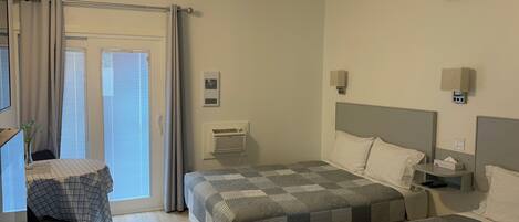 A hotel room with two beds, a desk, a chair, a window with curtains, an air conditioning unit, and a bedside table with a lamp.