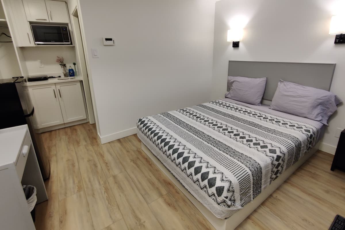 A compact bedroom with a bed, a nightstand, a wall-mounted light, and a kitchenette area with a microwave.