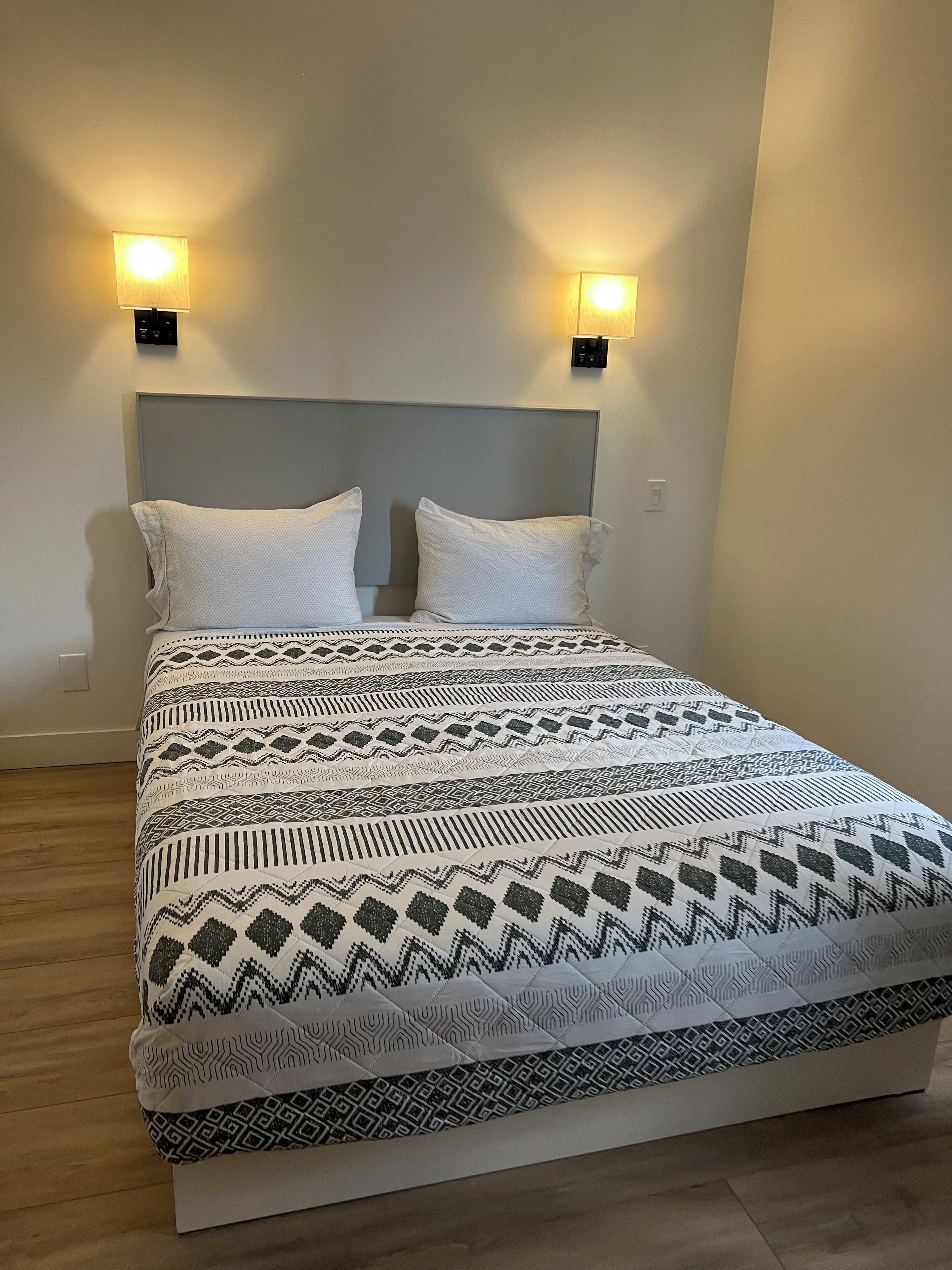A bed with a patterned bedspread, two wall-mounted lights, and a plain headboard.