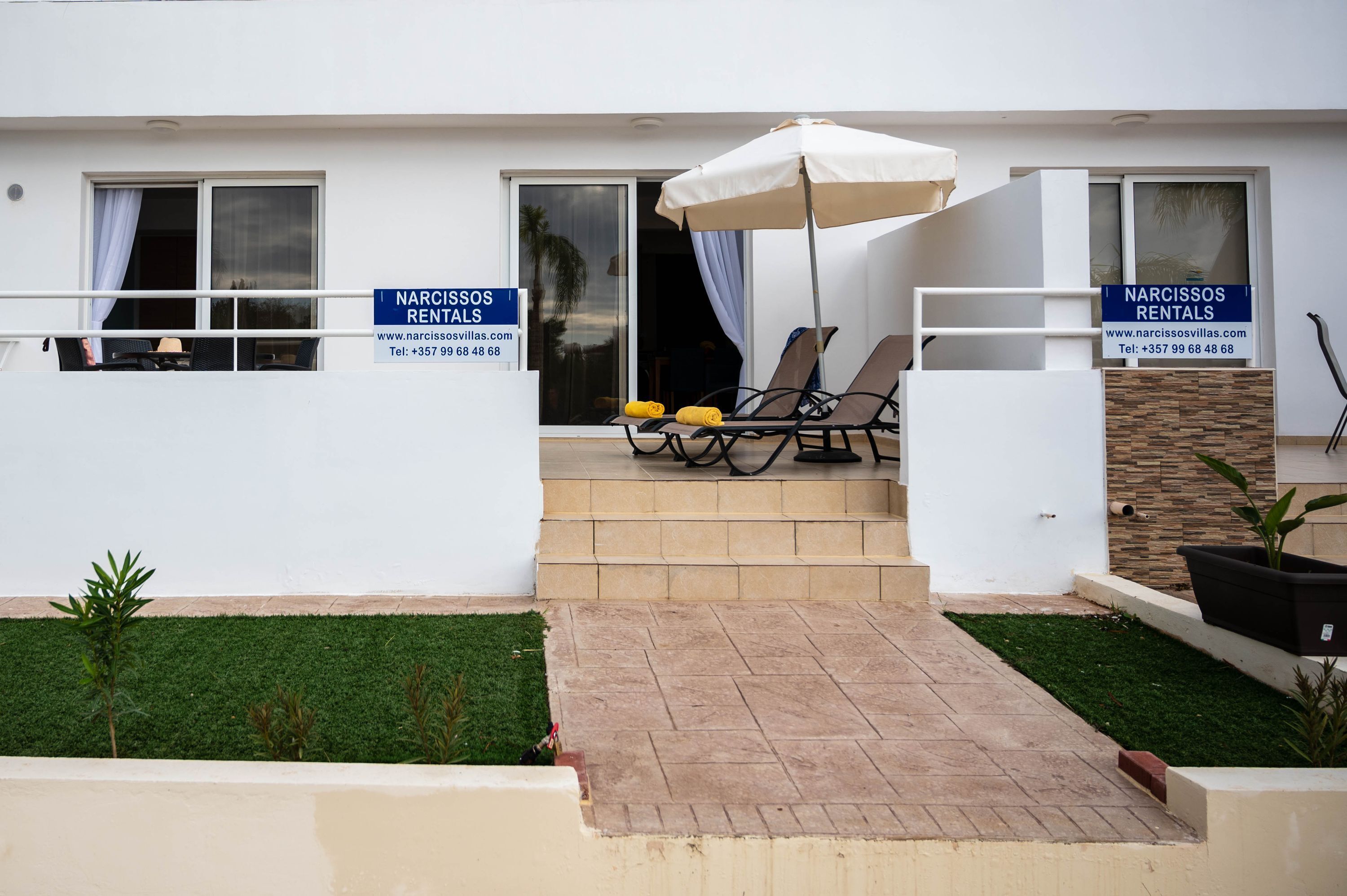 Une villa moderne dotée d’un patio comprenant deux chaises longues et un grand parasol. On y trouve des panneaux indiquant « NARCISSOS RENTALS » ainsi qu’un numéro de téléphone.