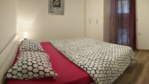 A bedroom with a bed, red bedding, a nightstand, a wall-mounted lamp, and a window with curtains. - Superior Apartment Tivat (Gošići)