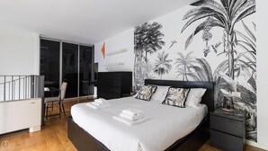 A bedroom with a large bed, a nightstand, a wall mural of tropical plants, a sliding door, and a small seating area with a table and chairs. - Italianway Comfort - Salomone 51 (Milan)