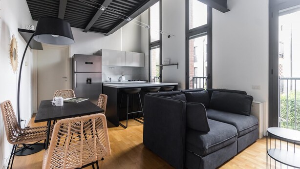A modern kitchen with a built-in refrigerator, a dining area with wicker chairs, and a dark grey sofa. - Italianway Comfort - Salomone 51 (Milan)