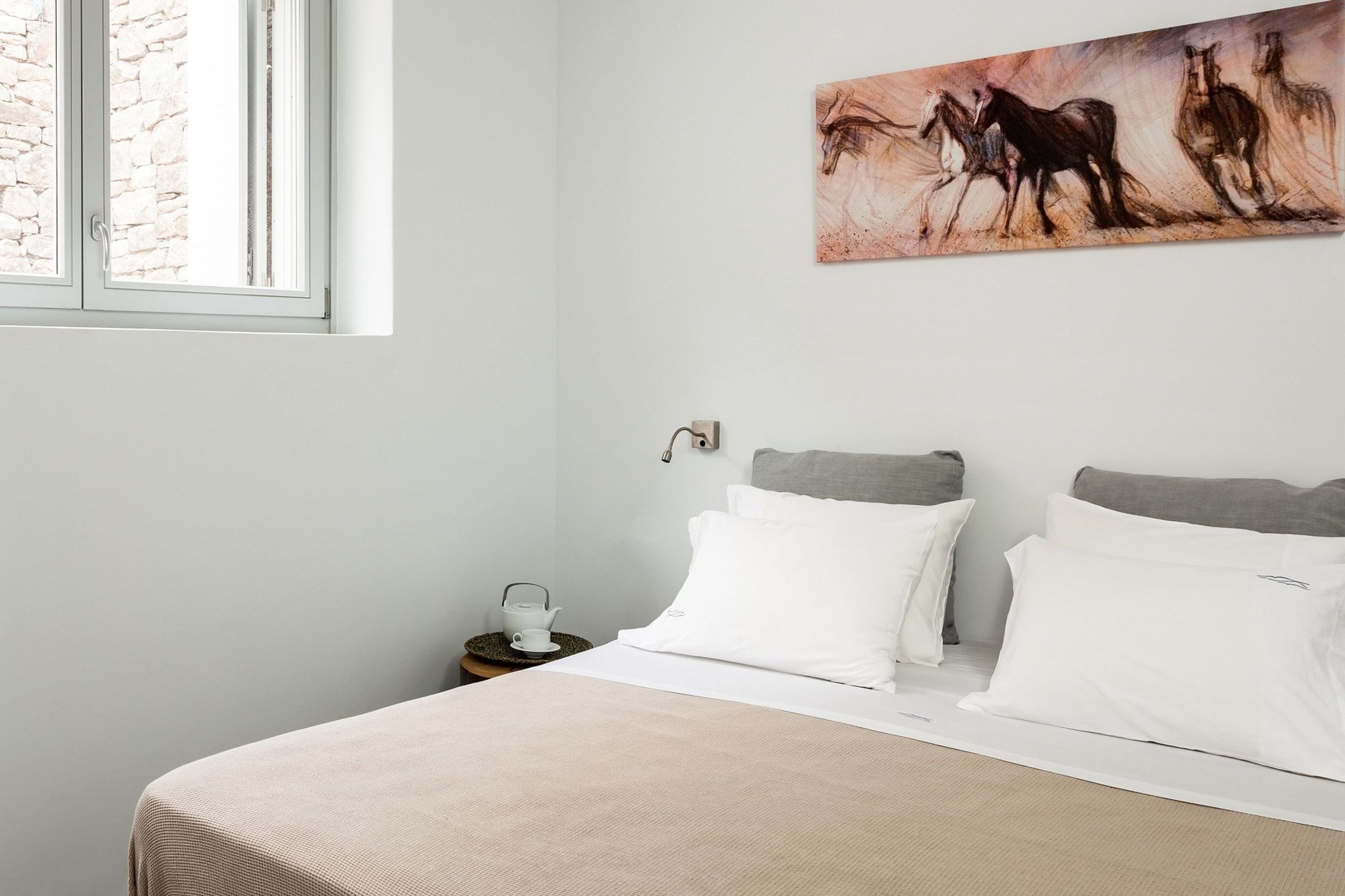 A bedroom with a bed, white pillows, a nightstand with a teapot, and a painting of horses on the wall.