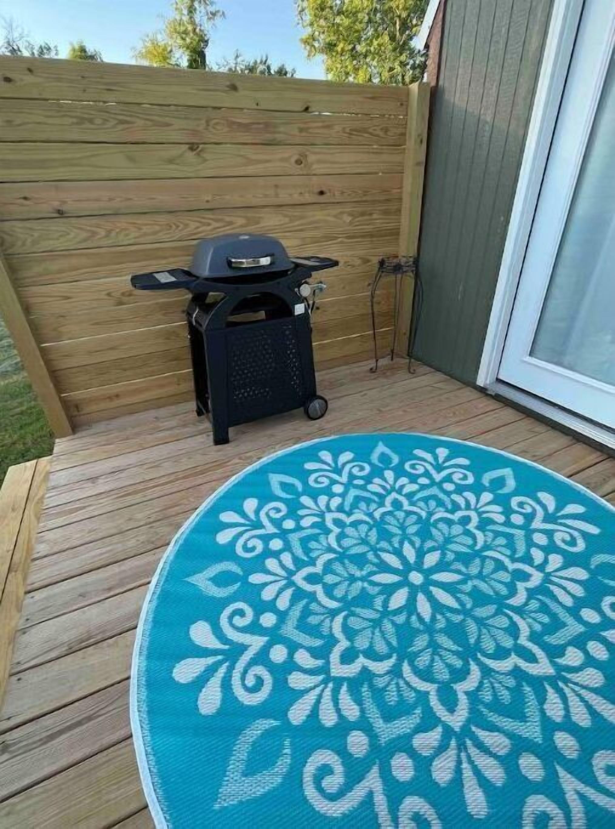 A wooden deck with a round blue patterned rug, a barbecue grill, and a sliding glass door.
