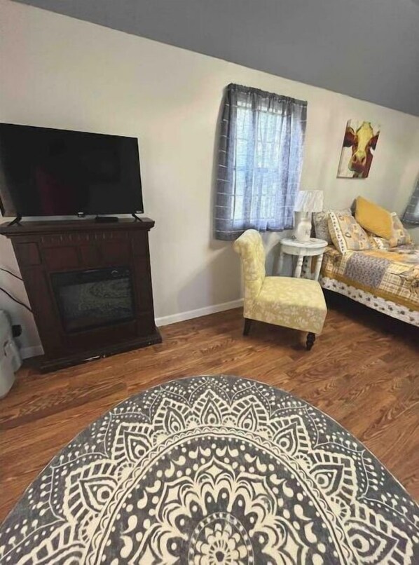Une pièce avec un téléviseur posé sur un meuble en bois, un tapis à motifs, un fauteuil jaune, un lit avec une literie à motifs et une petite table avec une lampe.