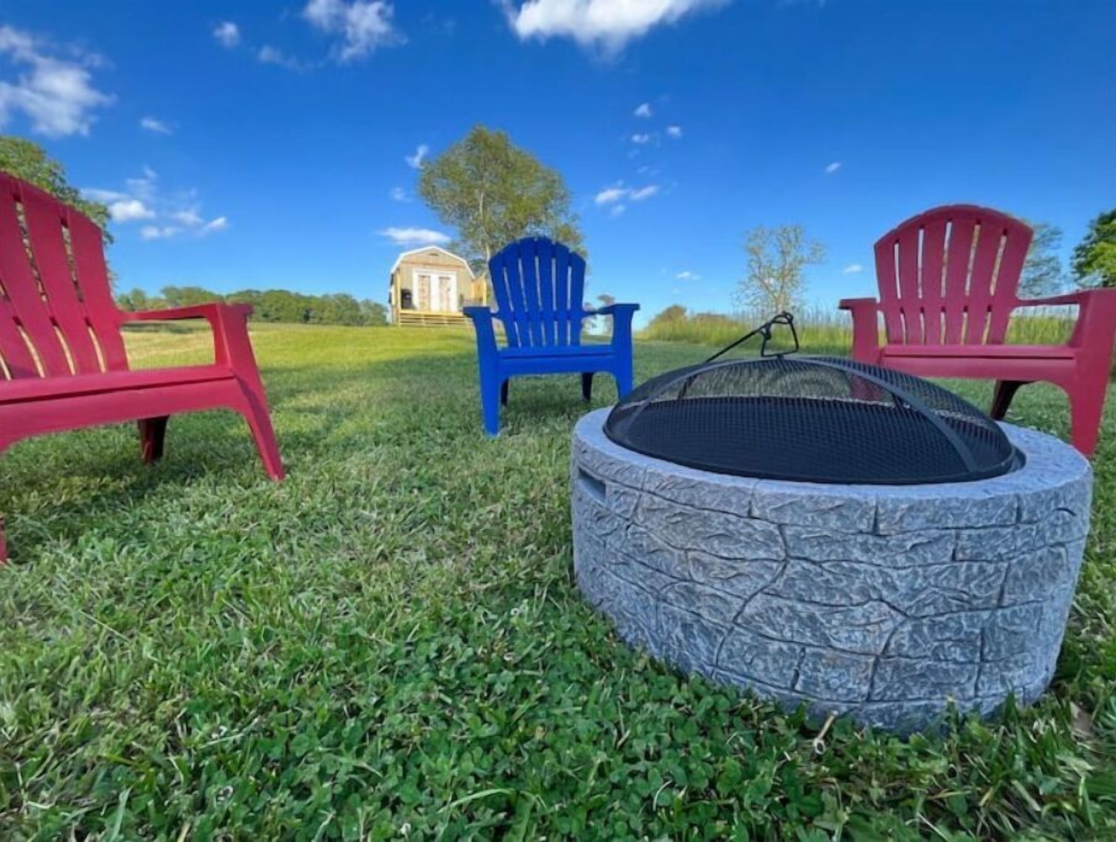 A circular stone fire pit with a metal grate, surrounded by red and blue Adirondack chairs, set on a grassy lawn with a clear blue sky and a distant building.