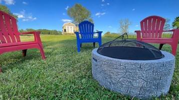 A circular stone fire pit with a metal grate, surrounded by red and blue Adirondack chairs, set on a grassy lawn with a clear blue sky and a distant building.