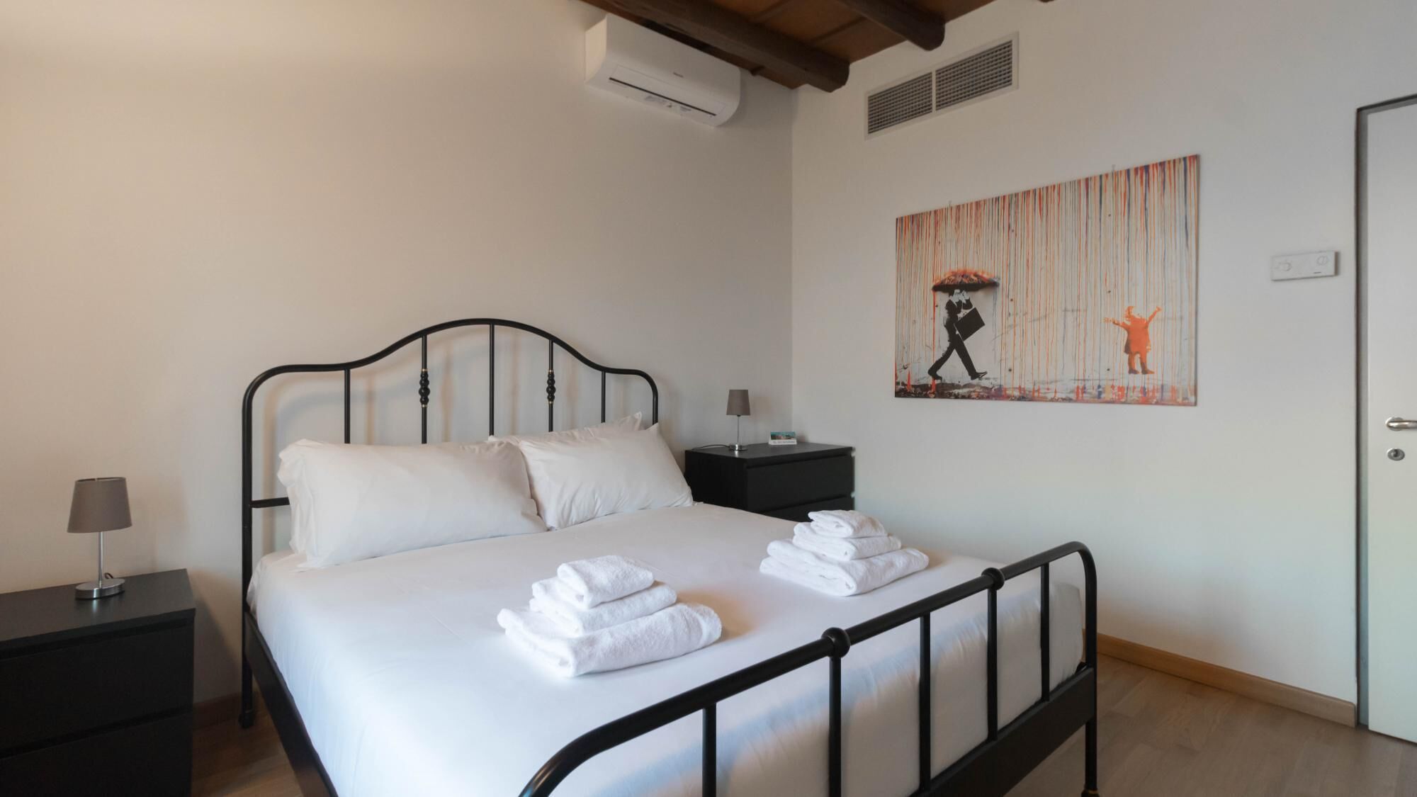 A neatly made bed with white linens and folded towels, a bedside table with a lamp, and a painting on the wall.