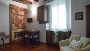 A cozy living room with a wooden bookshelf, a dining table with chairs, a radiator, and a window with sheer curtains. - Italianway Easy - San Calocero 29 (Milan)