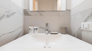 A modern bathroom with a large white sink, a mirror above it, and marble walls. There are toiletries on the sink and a light switch on the wall. - Italianway Easy - Ripa Ticinese 17 B (Milan)