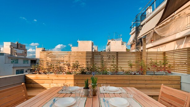 A rooftop terrace with a wooden table set for two, surrounded by potted plants and a wooden fence. - Premium Deluxe Apartment -c2-common Terrace (Piraeus)