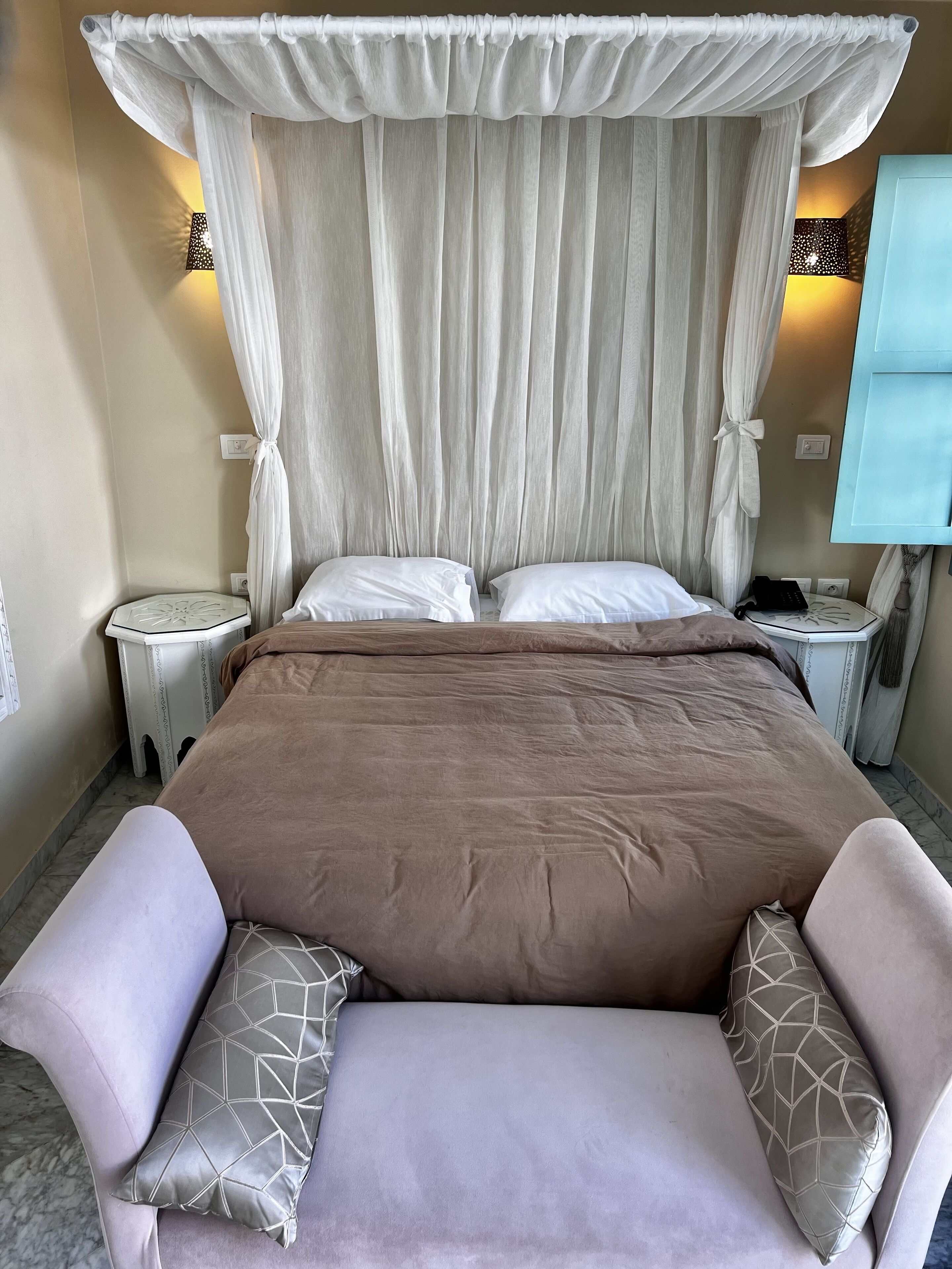 A neatly made bed with a brown bedspread, two pillows, and a canopy. There are two bedside tables with lamps, a wall-mounted light, and a blue cabinet.