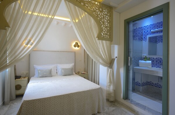 A bedroom with a large bed, a nightstand, a wall-mounted lamp, and a bathroom with blue tiles. - Dar Hammamet Guest House & Hammam (Hammamet)