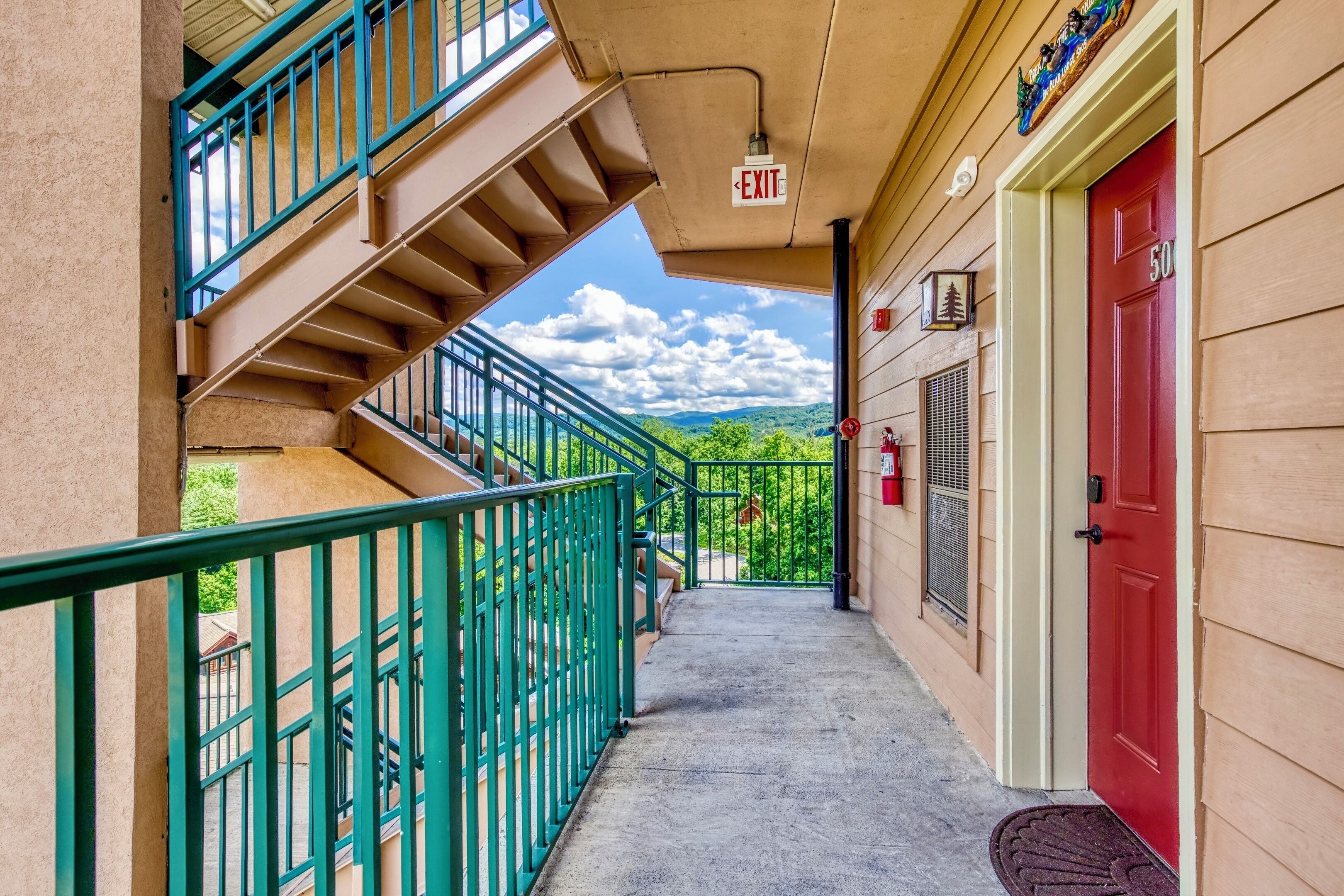 Un couloir avec une porte rouge, un panneau indiquant la sortie et un escalier menant à un espace extérieur agrémenté de verdure et offrant une vue sur la montagne.