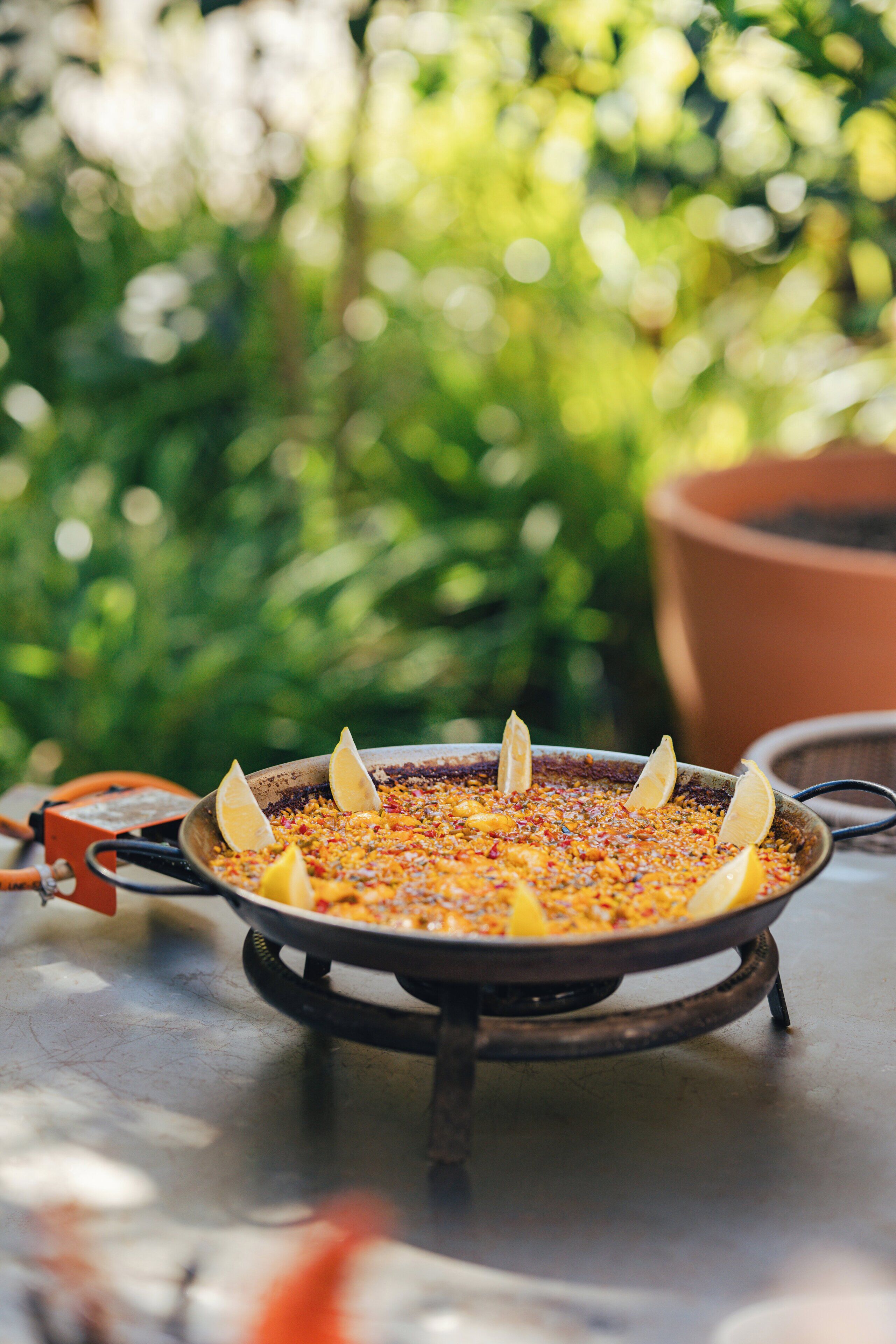 A pan of food on an outdoor stove with lemons on top, surrounded by greenery.
