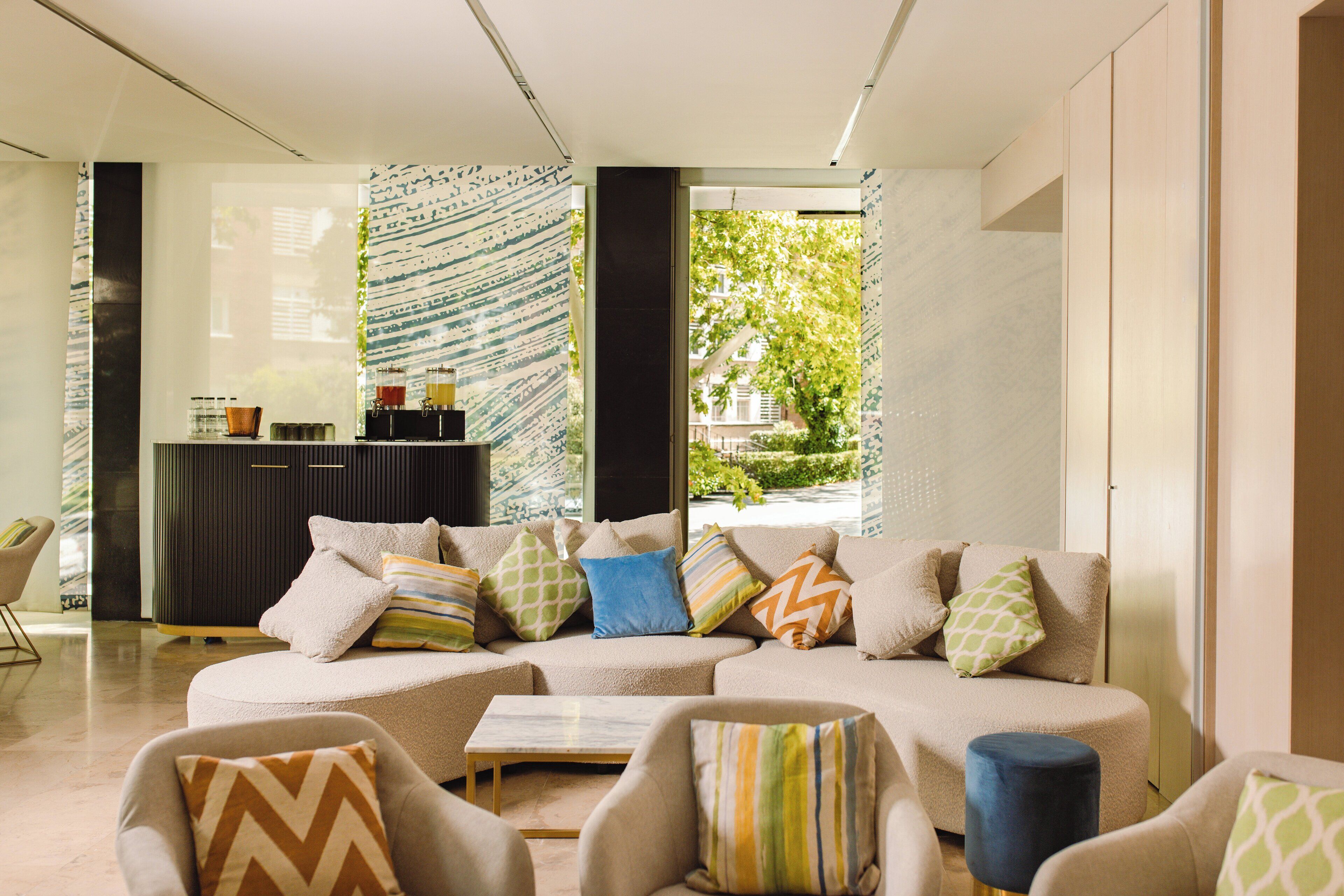 Een moderne woonkamer met een hoekbank, fauteuils en een salontafel. Er staat een donkere kast met decoratieve objecten en er is uitzicht op groen buiten.