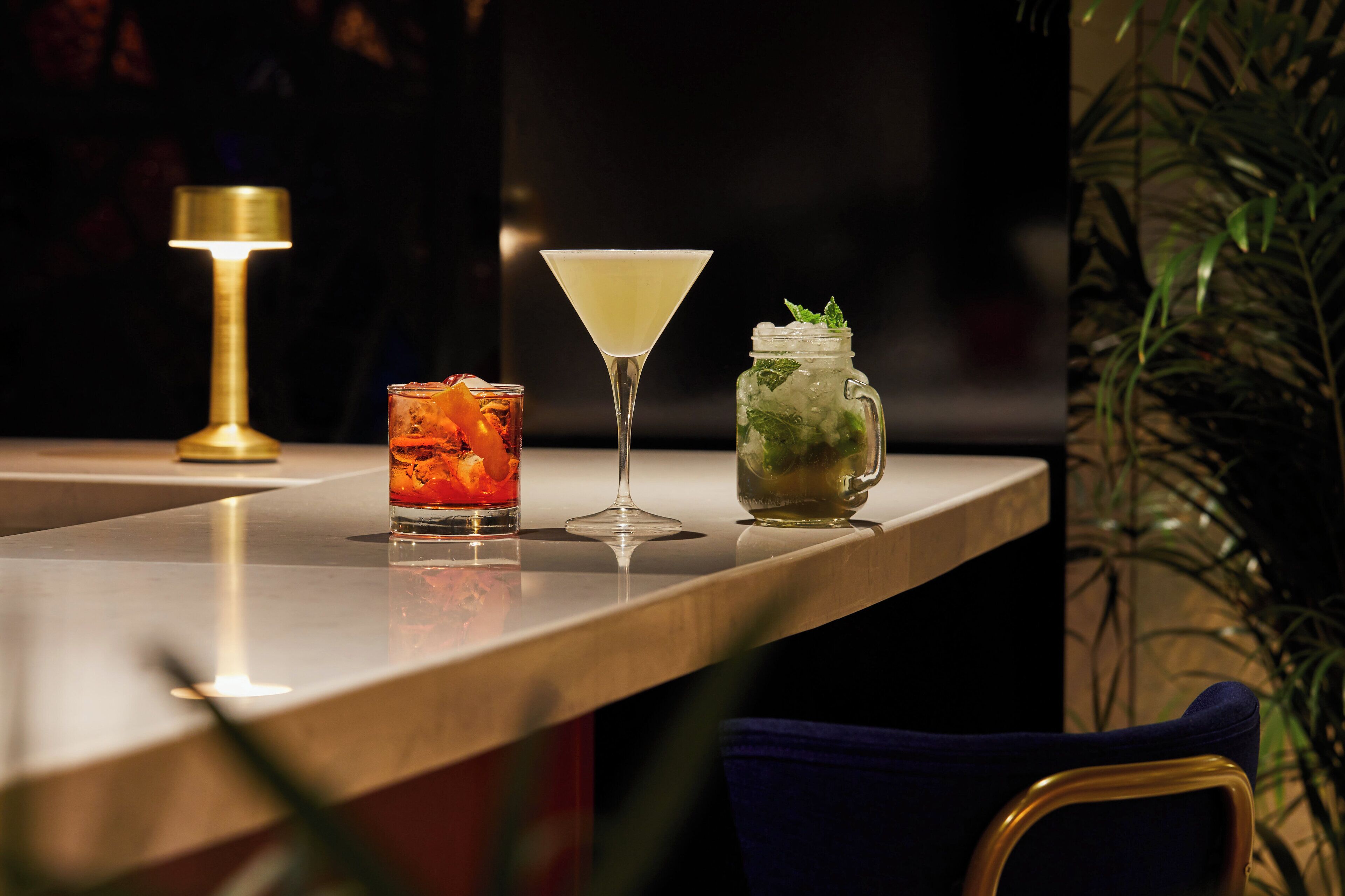 Eine Barbereich mit einem Martini-Glas, einem Cocktail mit Eis und Früchten sowie einem Einmachglas mit einem Getränk.
