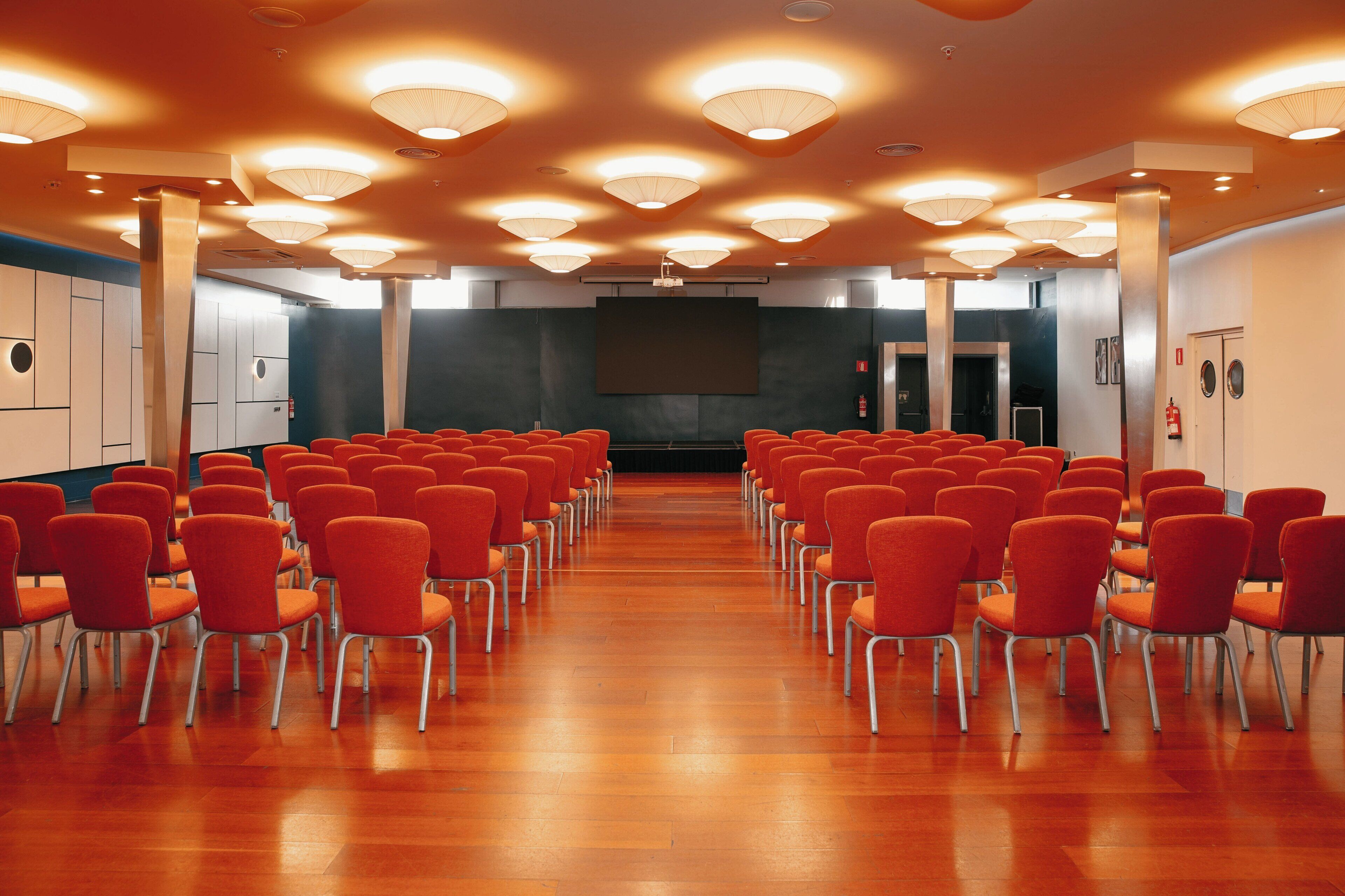 Une salle de conférence avec des rangées de chaises, un grand écran et des luminaires au plafond.