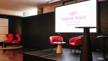 A modern hotel lobby with a large screen displaying the hotel's name and location. There are red chairs and a small table with a microphone.