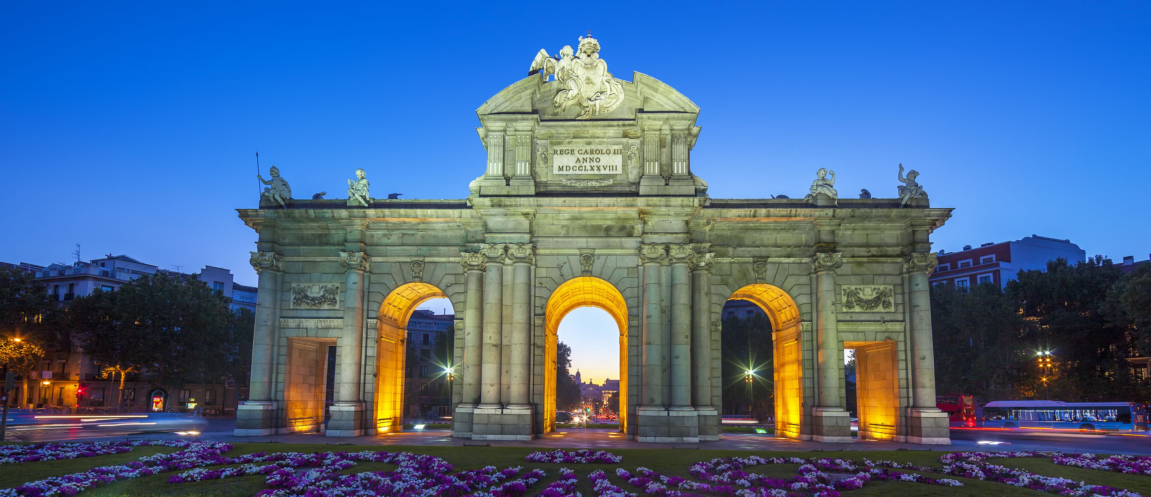Un arco monumental con inscripciones y estatuas, rodeado de un jardín bien cuidado con flores moradas.