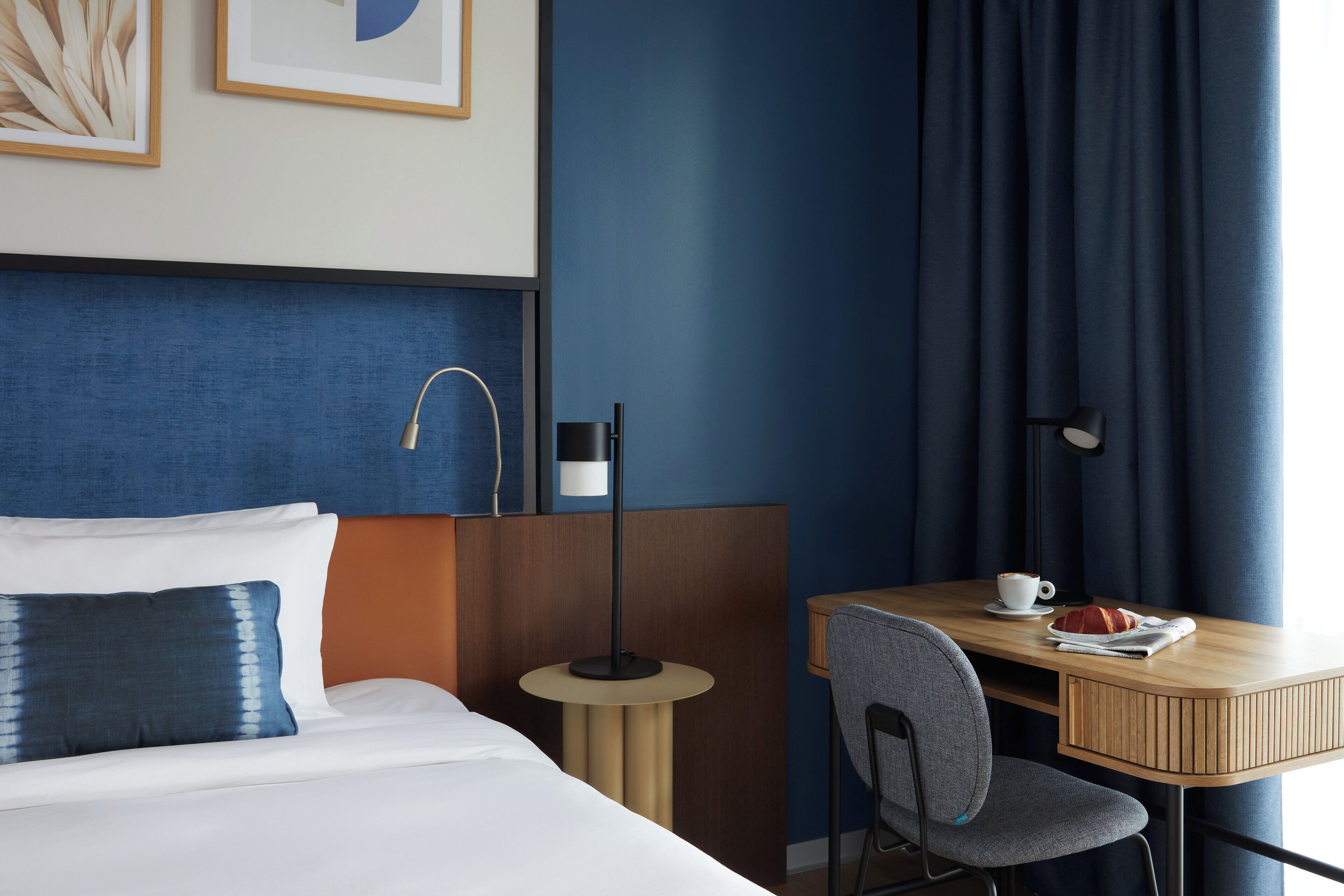A hotel room with a bed, a desk, a chair, a lamp, and a framed picture on the wall.