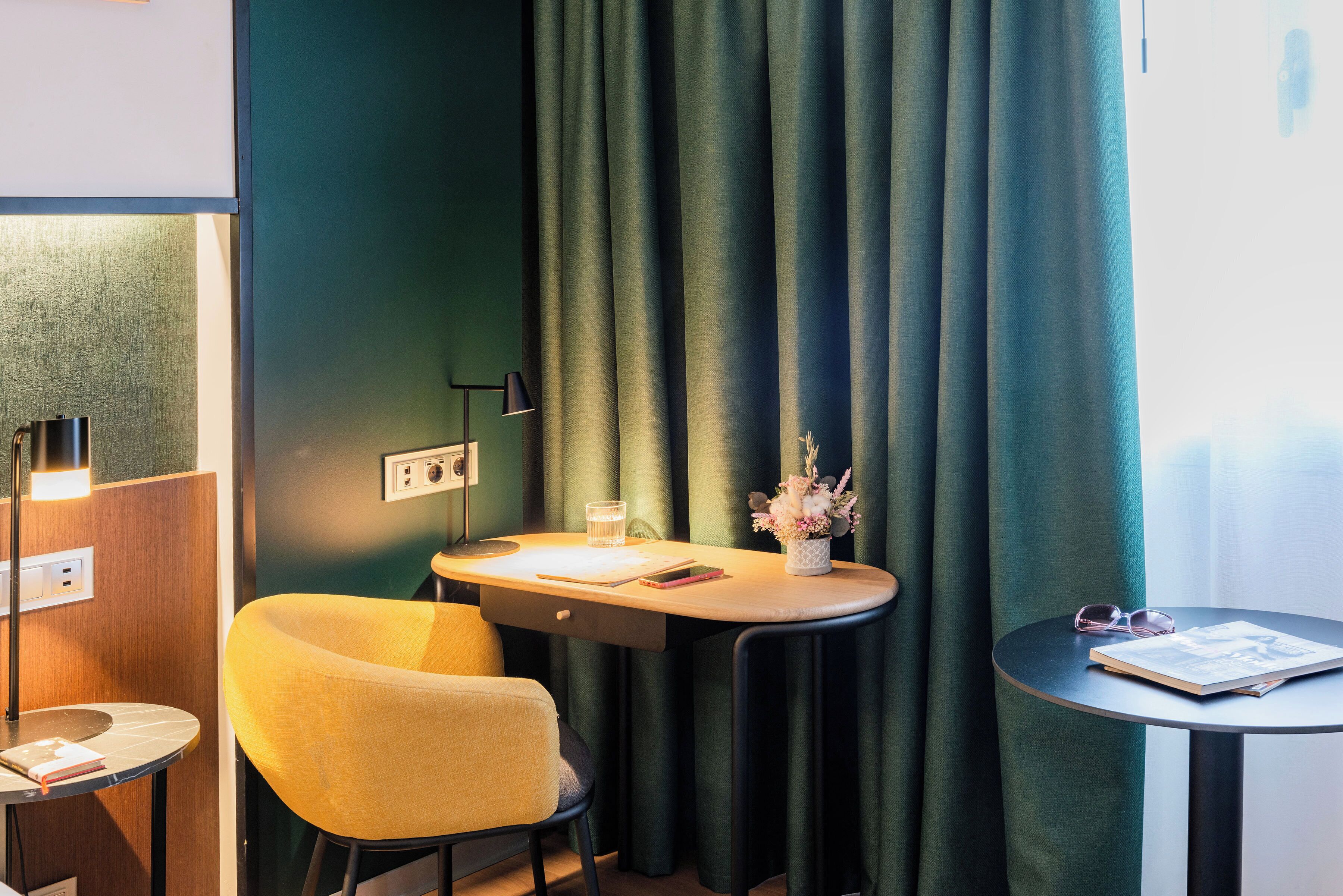 A room with a yellow chair, a small round table with a book and a glass, and a desk with a vase of flowers.
