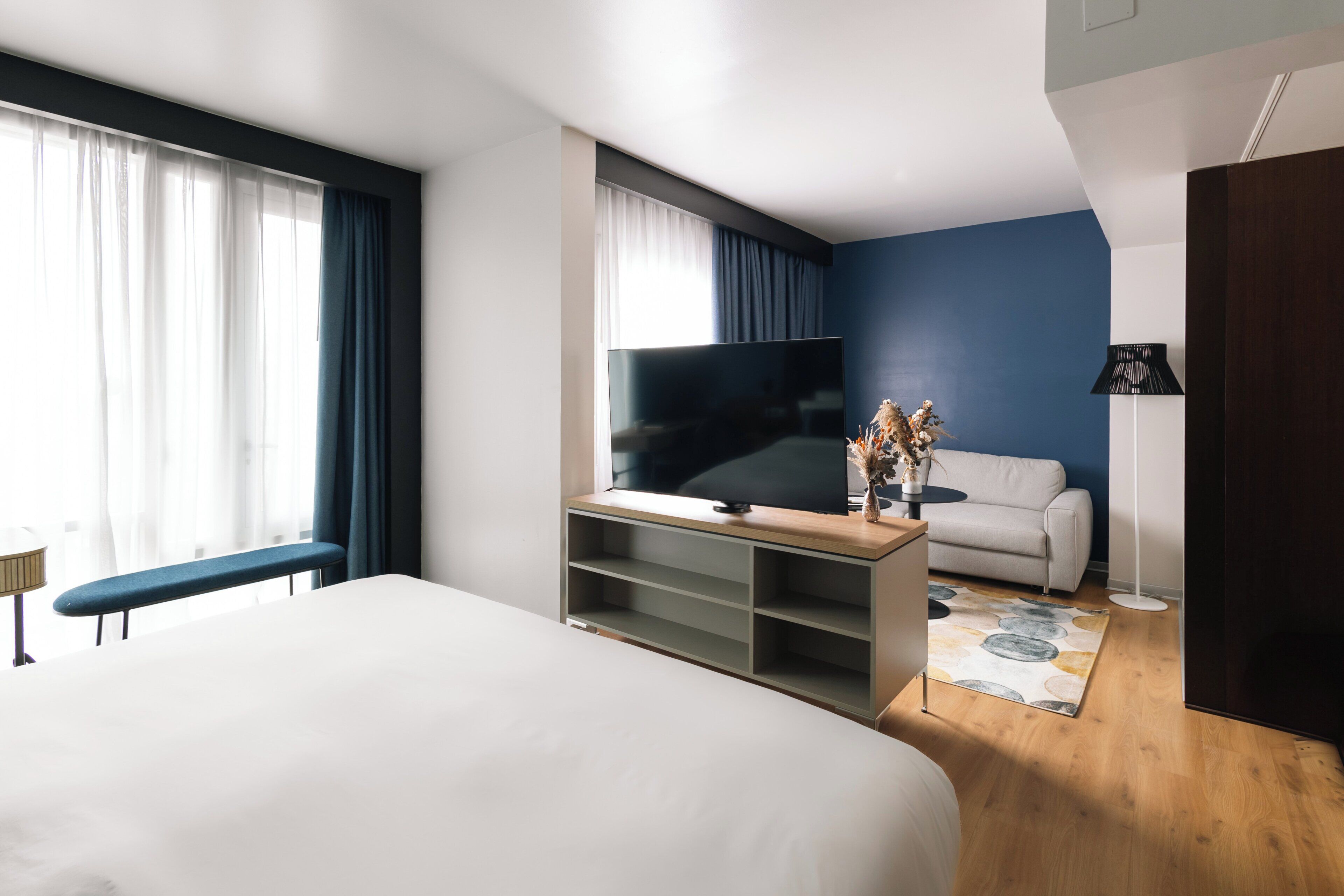 Una habitación de hotel moderna con una cama grande, un televisor de pantalla plana sobre un soporte de madera, un sofá y un escritorio con una lámpara.