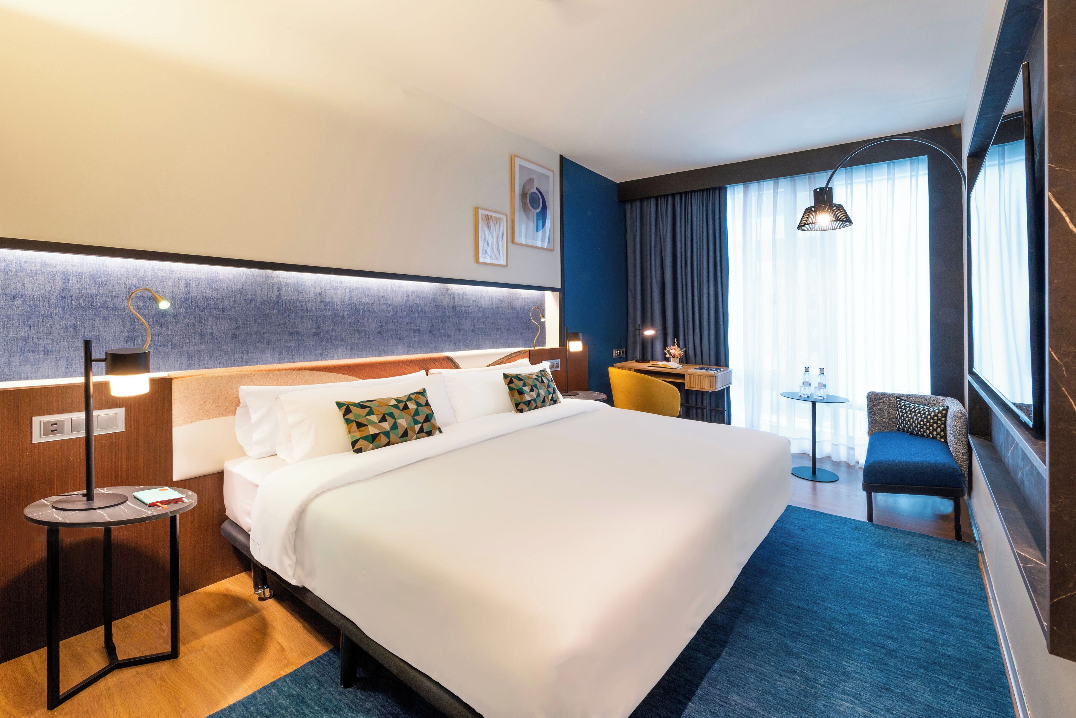 A hotel room with a large bed, a desk, a chair, and a small table.