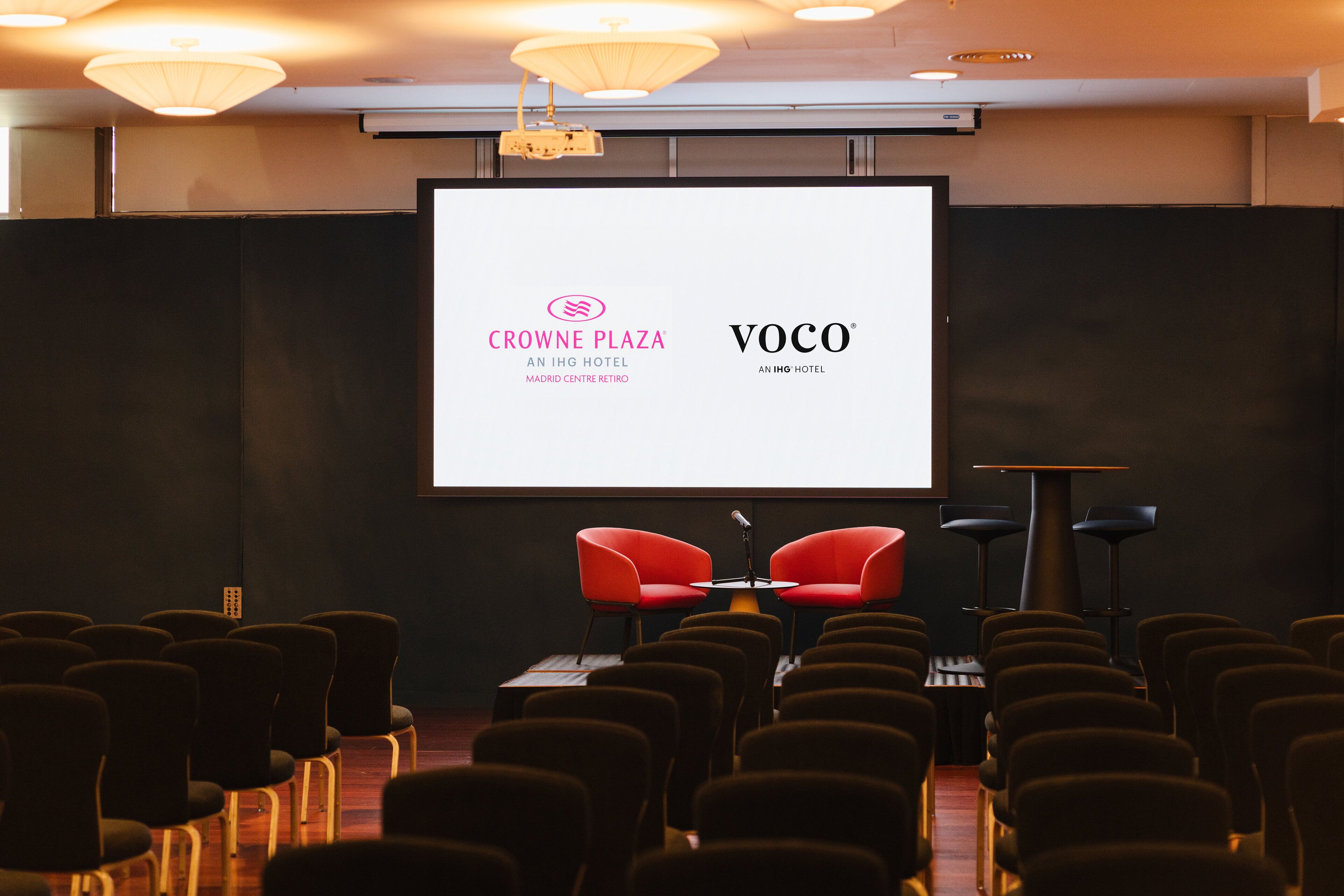 A conference room with rows of chairs, a large screen displaying logos, and two red chairs in the front.