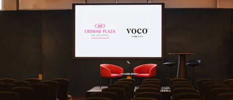 A conference room with rows of chairs, a large screen displaying logos, and two red chairs in the front.