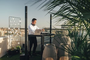 A person working on a laptop on a rooftop terrace with a city view, a decorative light fixture, and a wicker chair. - Crowne Plaza Madrid - Centre Retiro by IHG (Madrid)