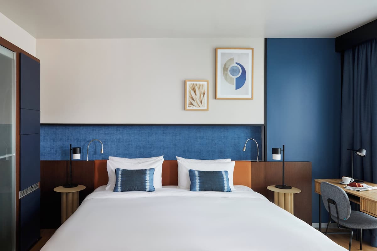 A hotel room with a large bed, two bedside tables, a desk with a chair, and artwork on the wall.