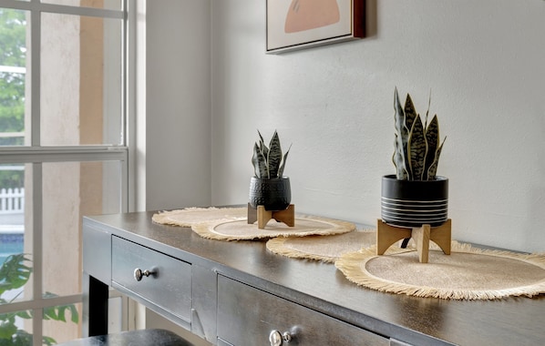 A dark wooden console table with two potted plants on woven placemats, a framed picture on the wall, and a window with a view of greenery. - Lakefront Screened Pool Home, Sleeps 12 | Sunset Cove Retreat, Riverview (Riverview)