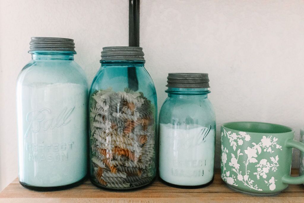 Three glass jars with lids, one containing pasta, another with a white powdery substance, and the third empty. A green floral mug is also visible.