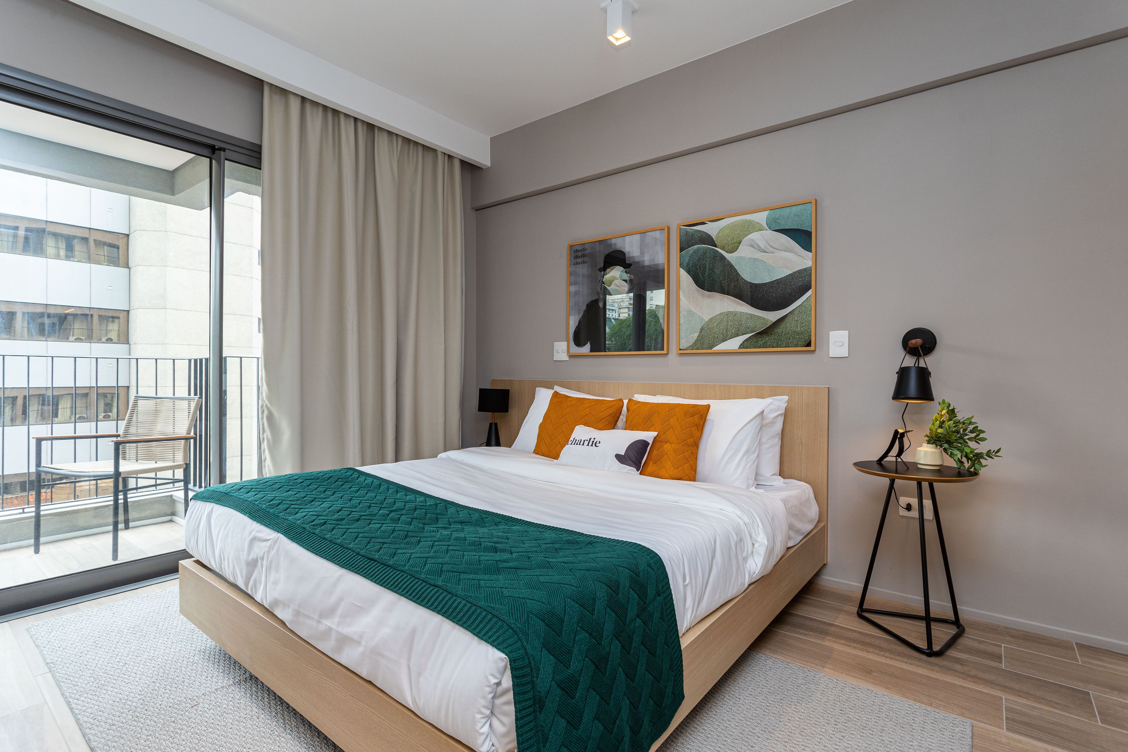 A modern hotel room with a large bed, a view of the city through floor-to-ceiling windows, a small side table with a lamp, and abstract wall art.
