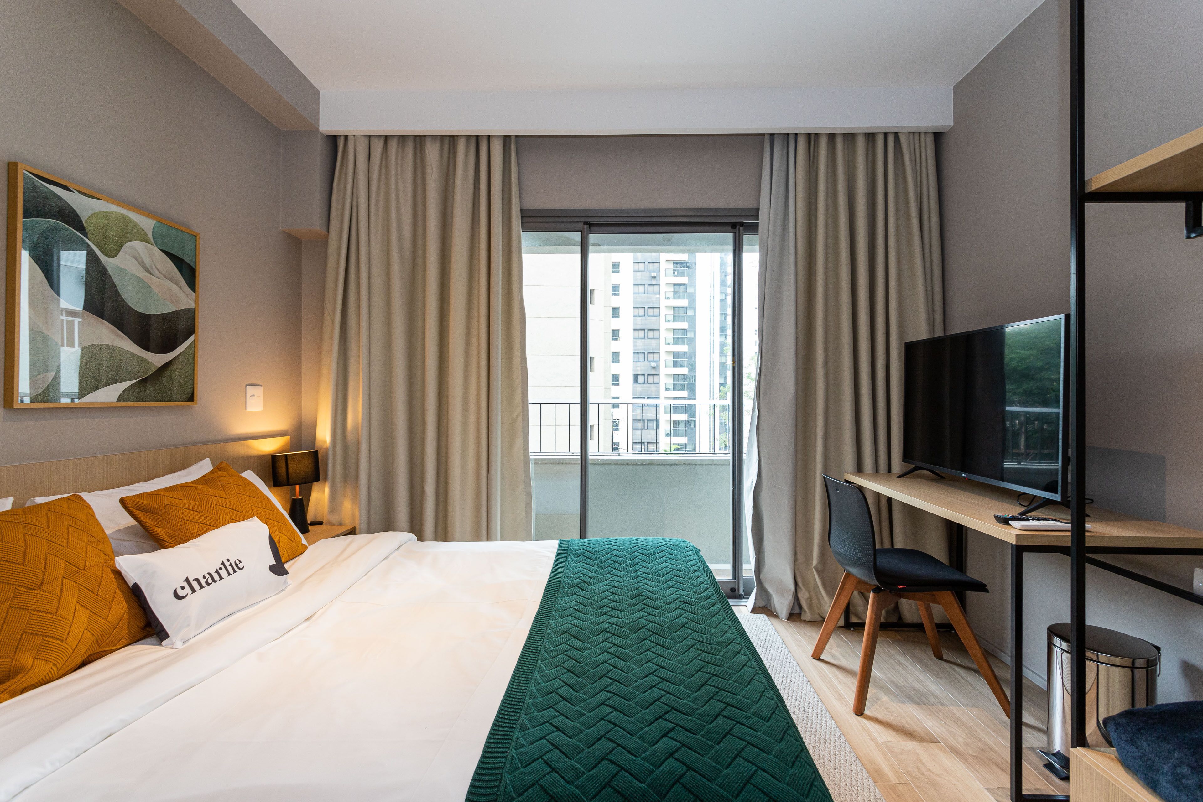 A hotel room with a bed, a desk with a TV, a window with curtains, and a painting on the wall.