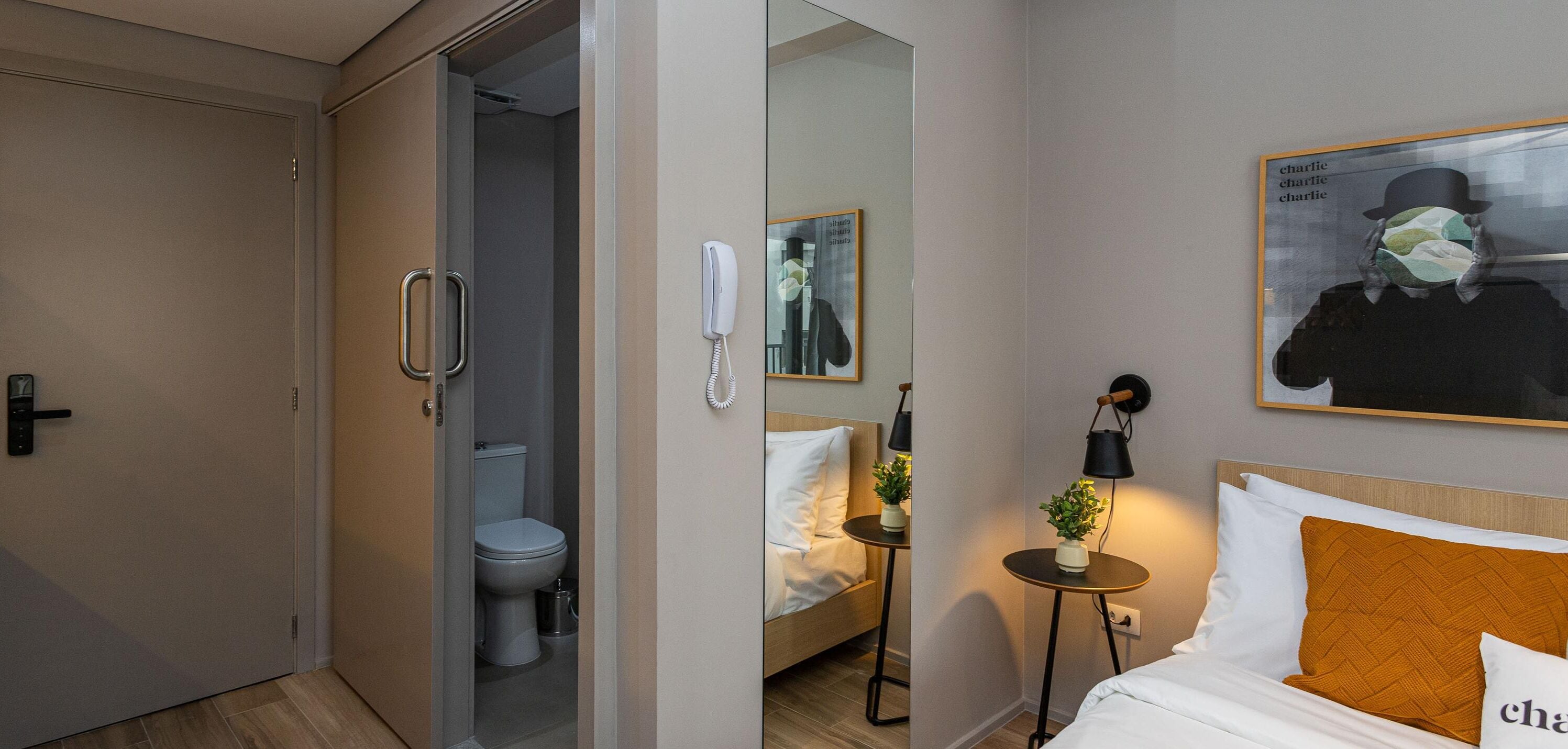 A hotel room with a bed, a bedside table with a lamp, a small potted plant, a wall-mounted phone, and a painting.