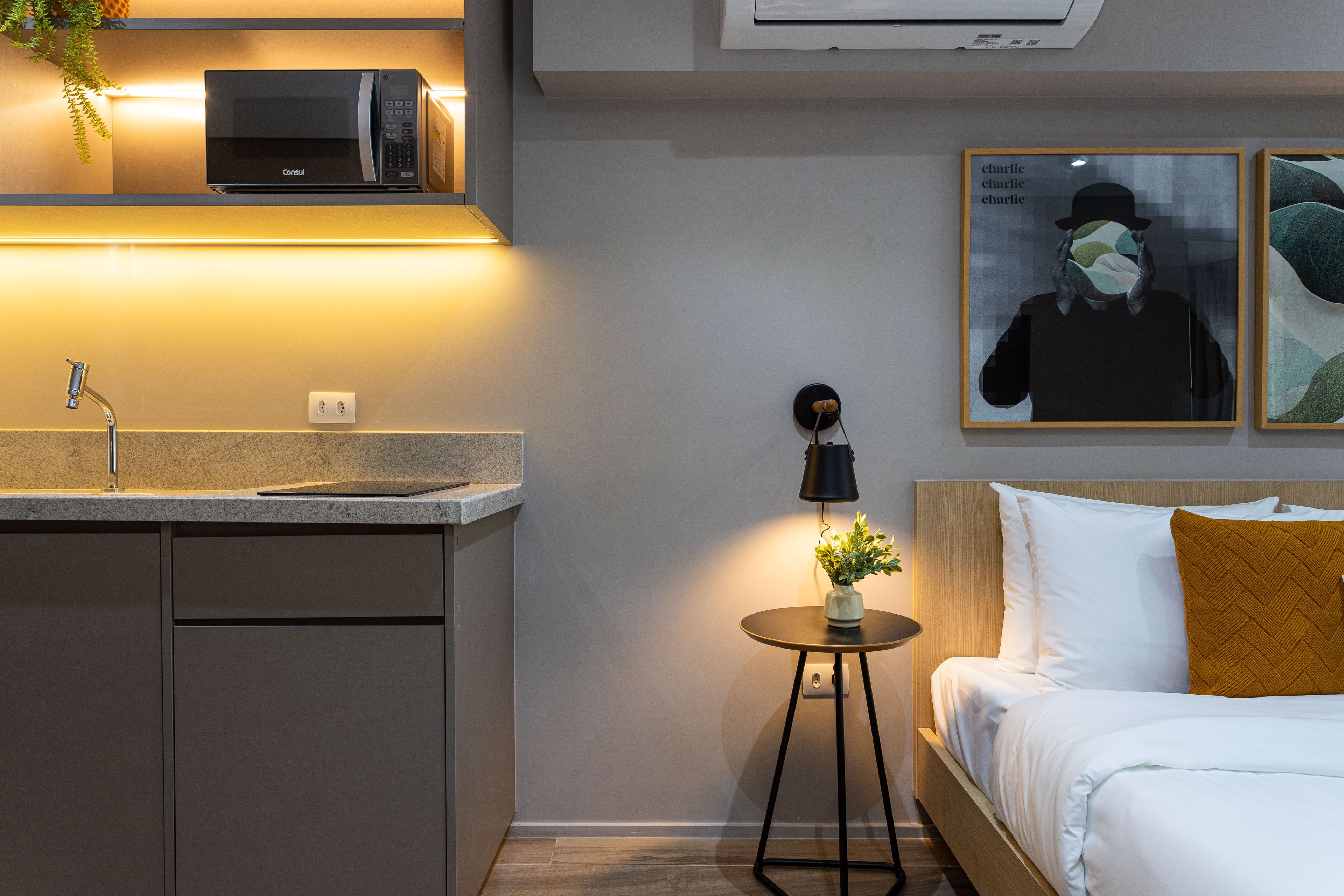 A modern hotel room with a kitchenette, a bedside table with a lamp, a bed with white bedding and a mustard-colored pillow, and two framed artworks on the wall.