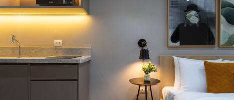 A modern hotel room with a kitchenette, a bedside table with a lamp, a bed with white bedding and a mustard-colored pillow, and two framed artworks on the wall.