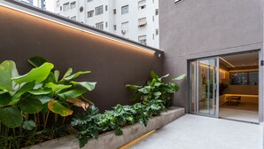 Un edificio moderno con facciata in cemento, un vaso rialzato pieno di piante verdi rigogliose, pavimento piastrellato e una porta in vetro che dà su un ambiente interno.